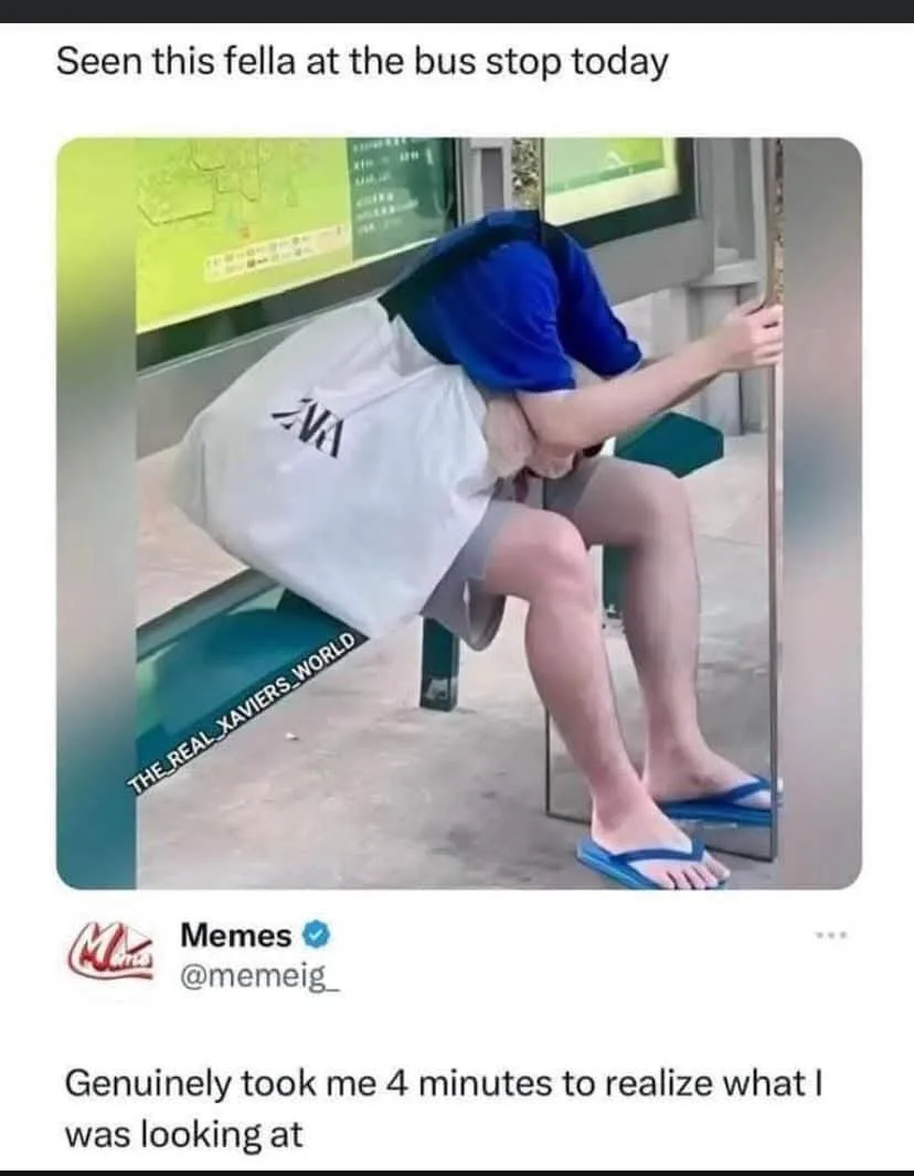 A person sits on a bus stop bench, leaning forward with their head down and arms wrapped around a large white tote bag. The…