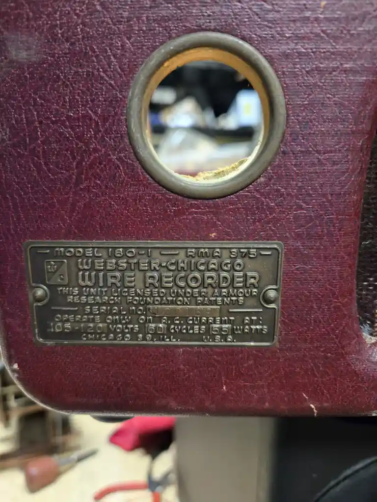 A close-up of a vintage maroon Webster-Chicago Wire Recorder with a brass plaque showing model, serial number, and operating…