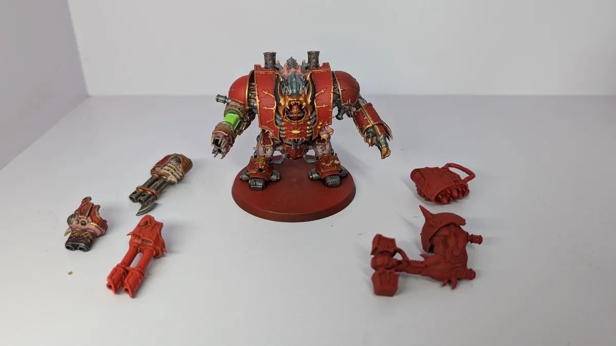 A heavily painted red and gold fantasy warlord miniature stands on a base, surrounded by several unpainted red plastic parts…