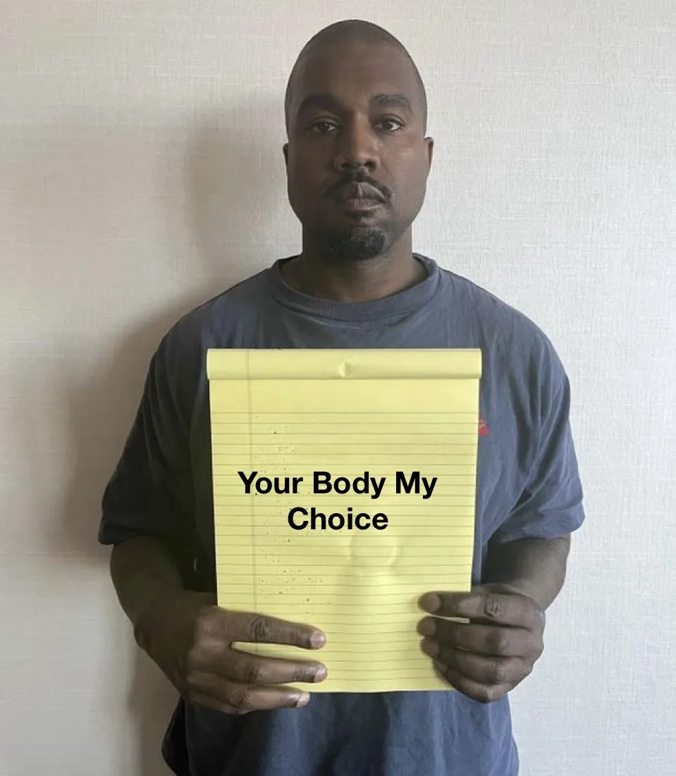 A man in a gray t-shirt stands against a plain wall holding a yellow legal pad. The pad has the text 'Your Body My Choice'…
