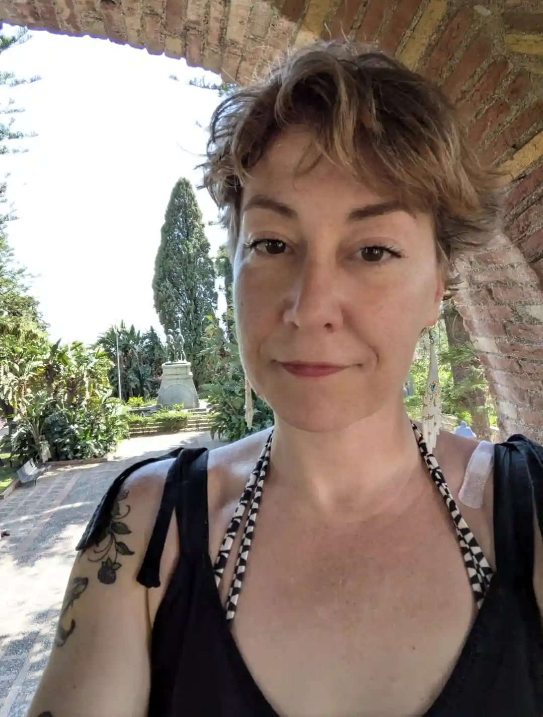 A woman with short brown hair and a tattoo on her left shoulder takes a selfie under a brick archway, smiling slightly while…