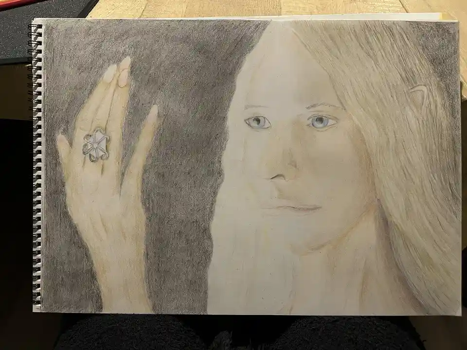 A colored pencil drawing in a spiral-bound sketchbook shows a person with long blonde hair and pointed ears, holding up a…