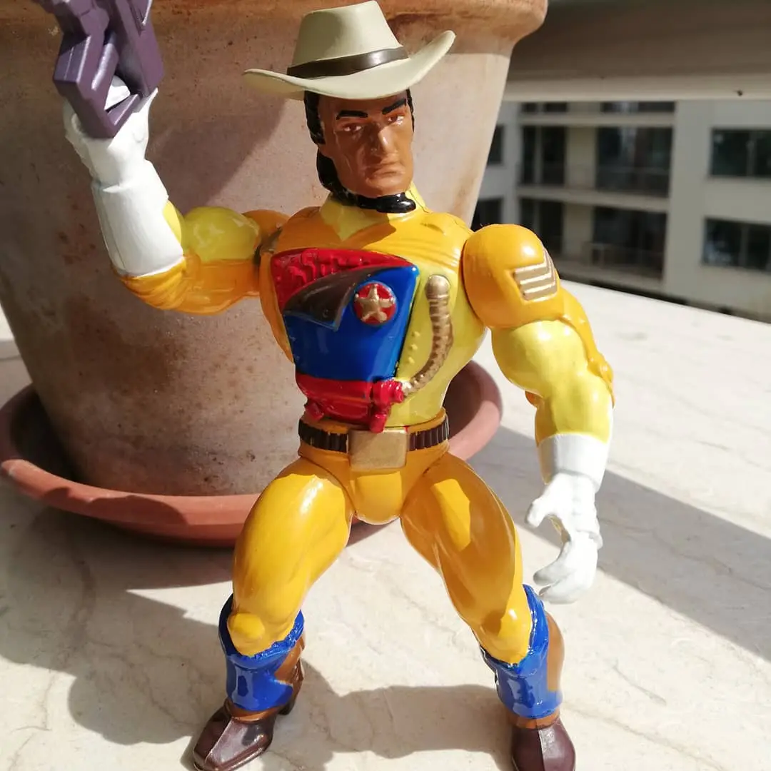 A vintage action figure of a muscular cowboy in a yellow suit, white hat, and blue boots, holding a purple gun, stands on a…