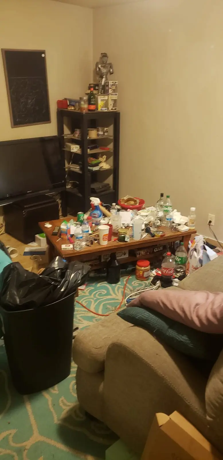 A messy living room with a coffee table overflowing with bottles, cups, and trash, a couch with pillows, a black trash can…