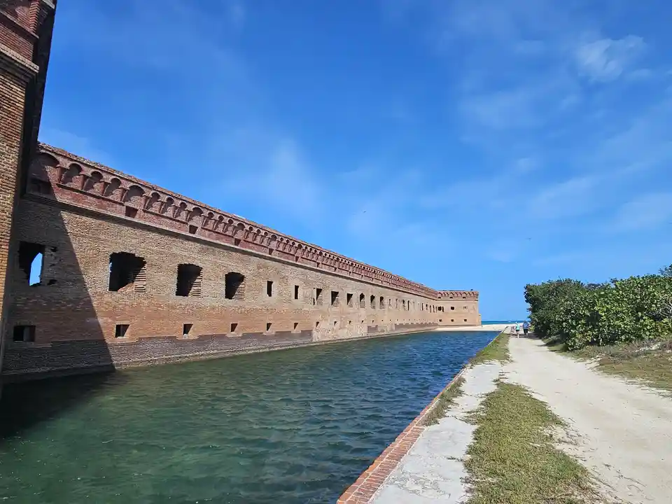 A long brick fortress wall runs alongside a narrow canal under a bright blue sky, with a sandy path and greenery on the…