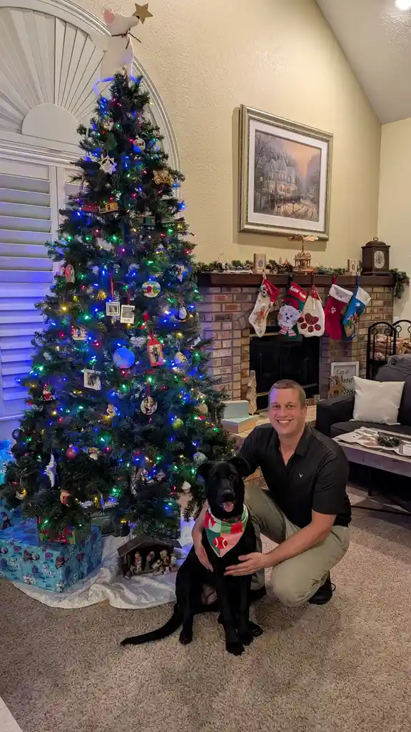 A man kneels beside a black dog wearing a festive bandana, posing in front of a brightly lit, heavily decorated christmas…