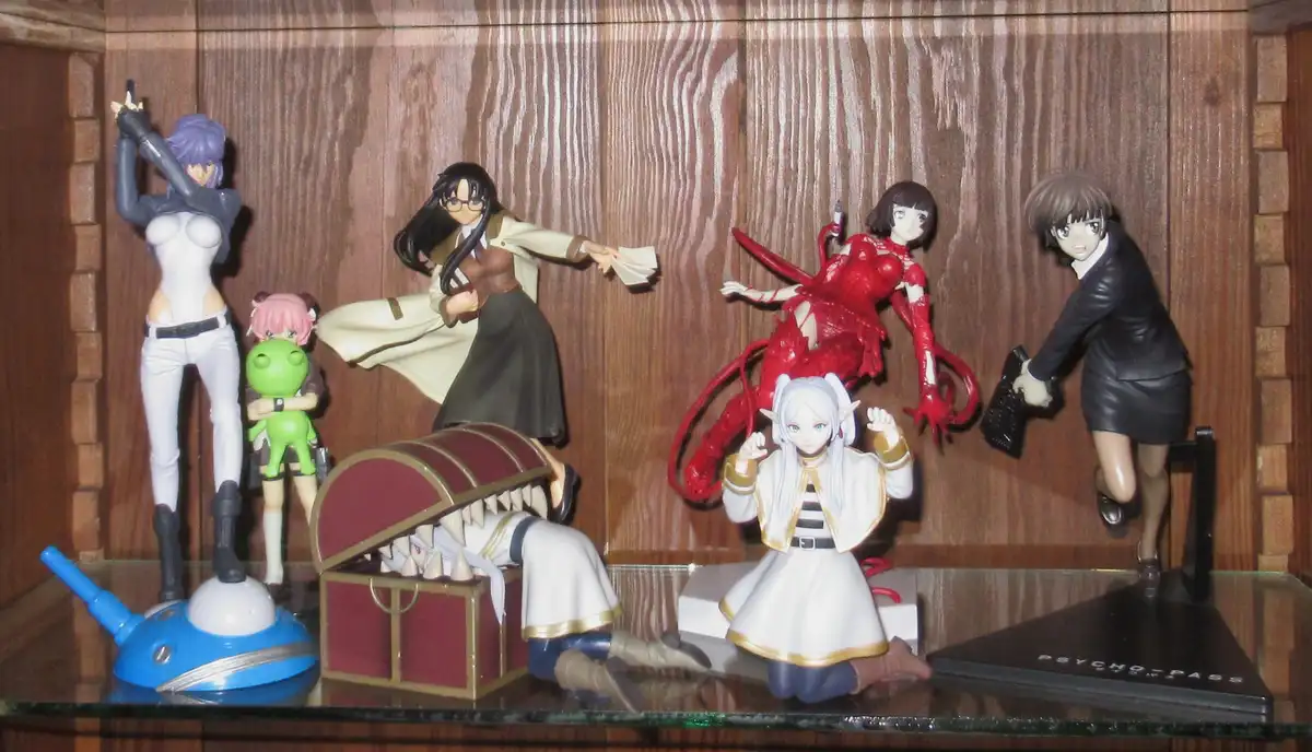 A collection of anime figurines, including characters from Psycho-Pass, are arranged on a glass shelf against a wooden…