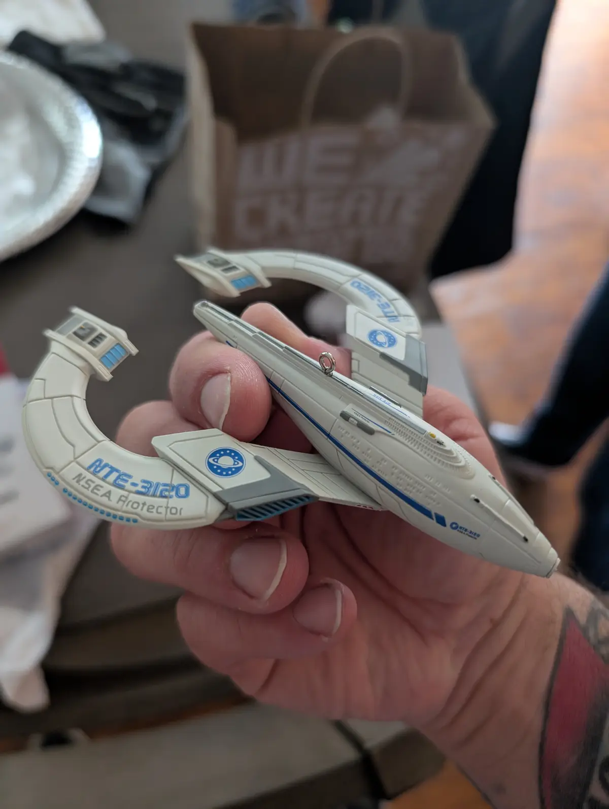 A hand holds a small, white and blue model of a spaceship labeled 'NTE-3120 NSEA Protector' with a ring for a keychain. The…