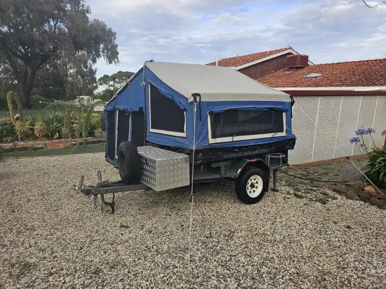A blue and grey pop-up camper trailer is parked on a gravel driveway in front of a house with a red-tiled roof and white…