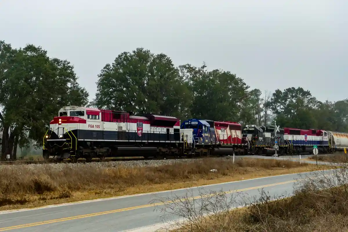 A long freight train with multiple locomotives in red, white, and blue livery is passing by a road with trees in the…