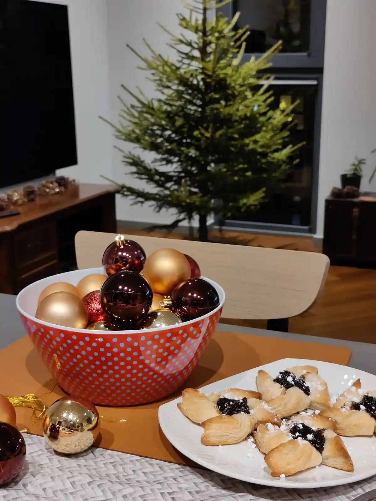 A festive indoor scene shows a red bowl with blue polka dots filled with shiny gold and burgundy Christmas ornaments, placed…