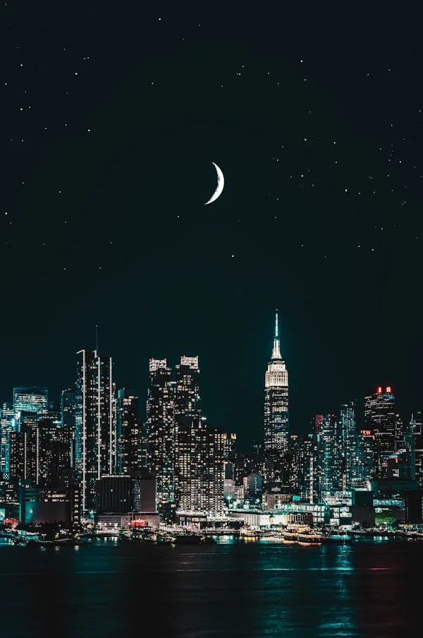 A nighttime cityscape featuring a crescent moon and stars above a lit-up skyline, with the Empire State Building prominently…