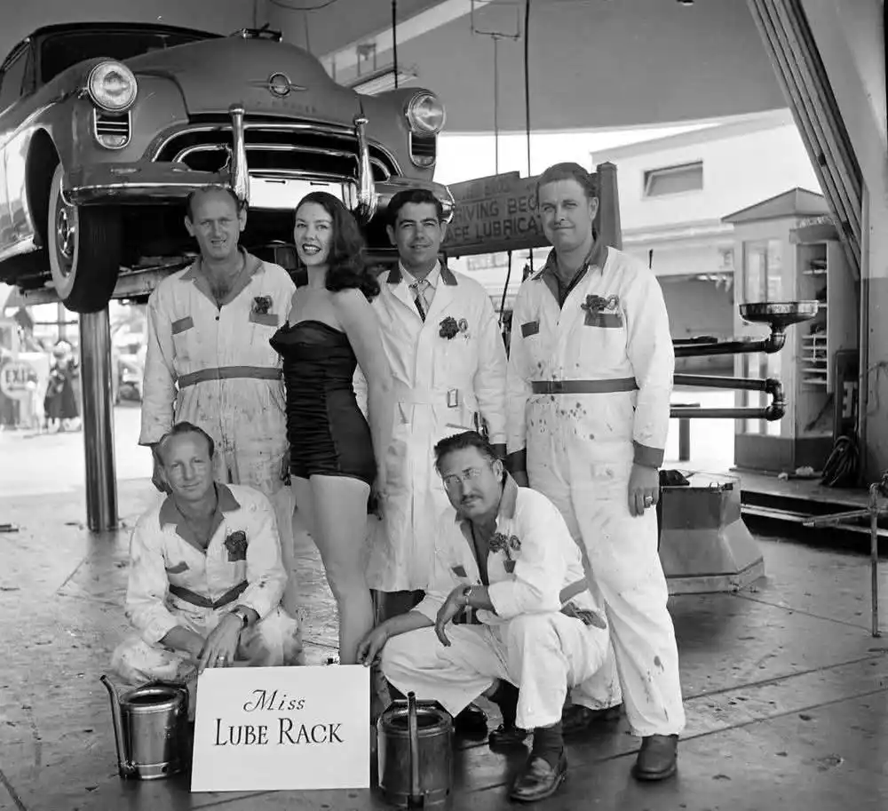 A vintage black-and-white photo shows six people—five men in mechanic overalls and one woman in a black strapless…