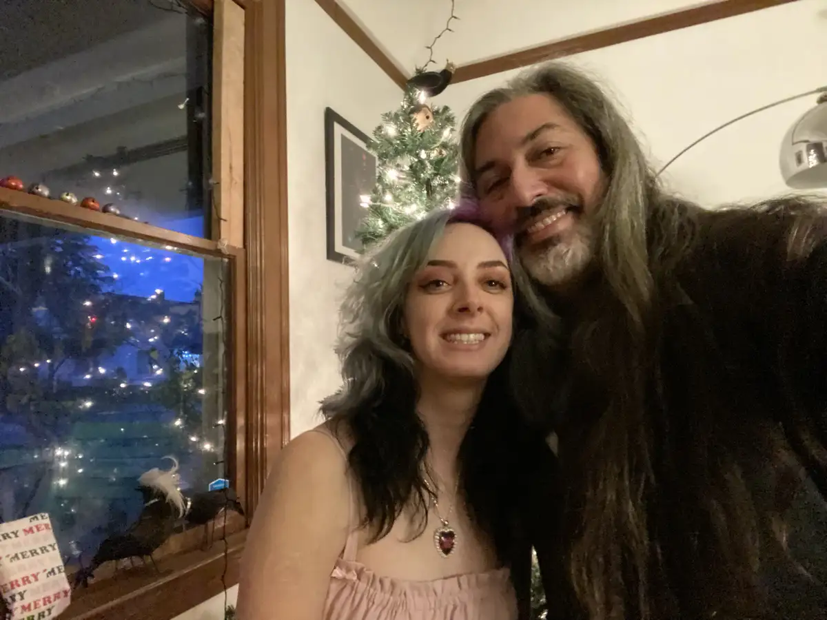 A selfie of a smiling man with long gray hair and a beard and a woman with blue-green hair, both smiling and posing closely…