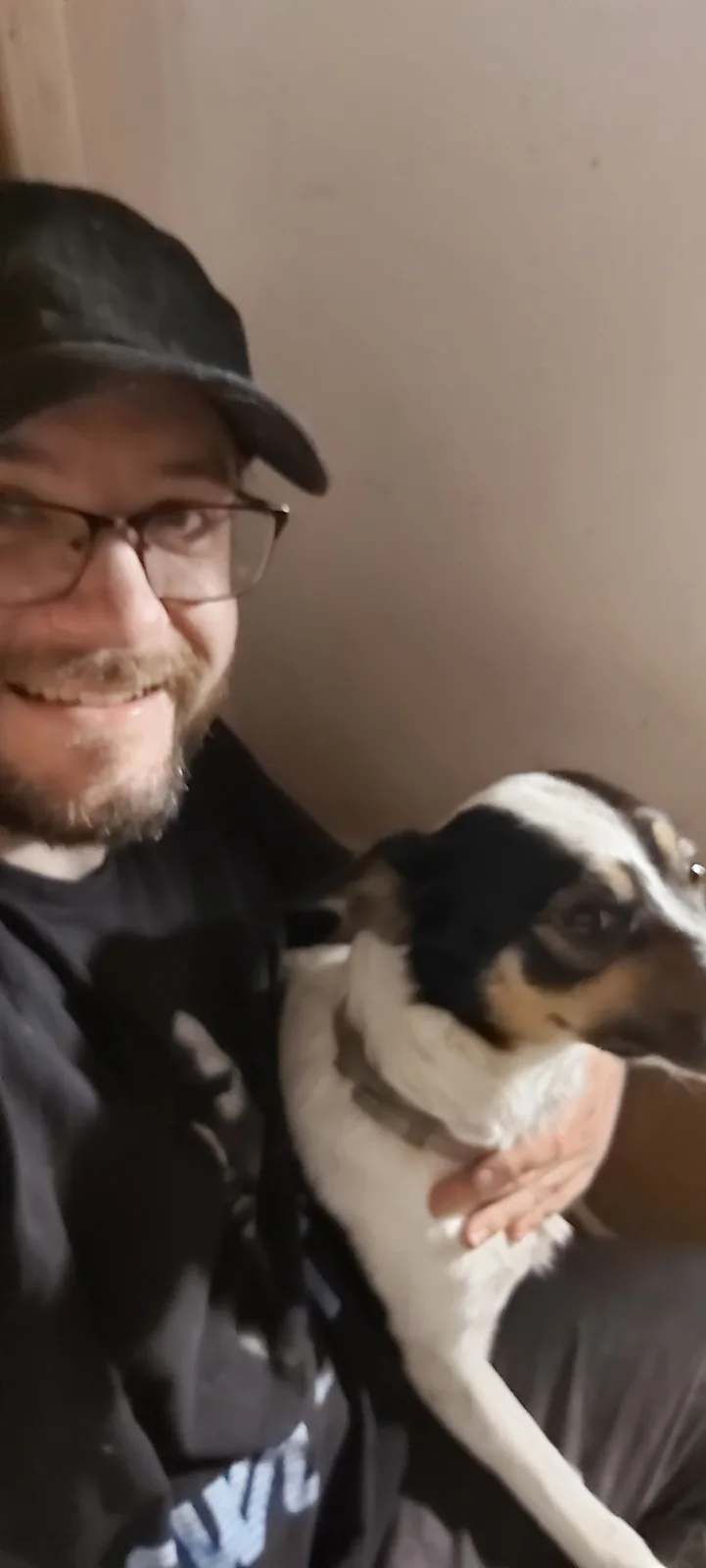 A blurry selfie shows a man with glasses and a cap smiling while holding a black and white dog. The dog is nestled in his…