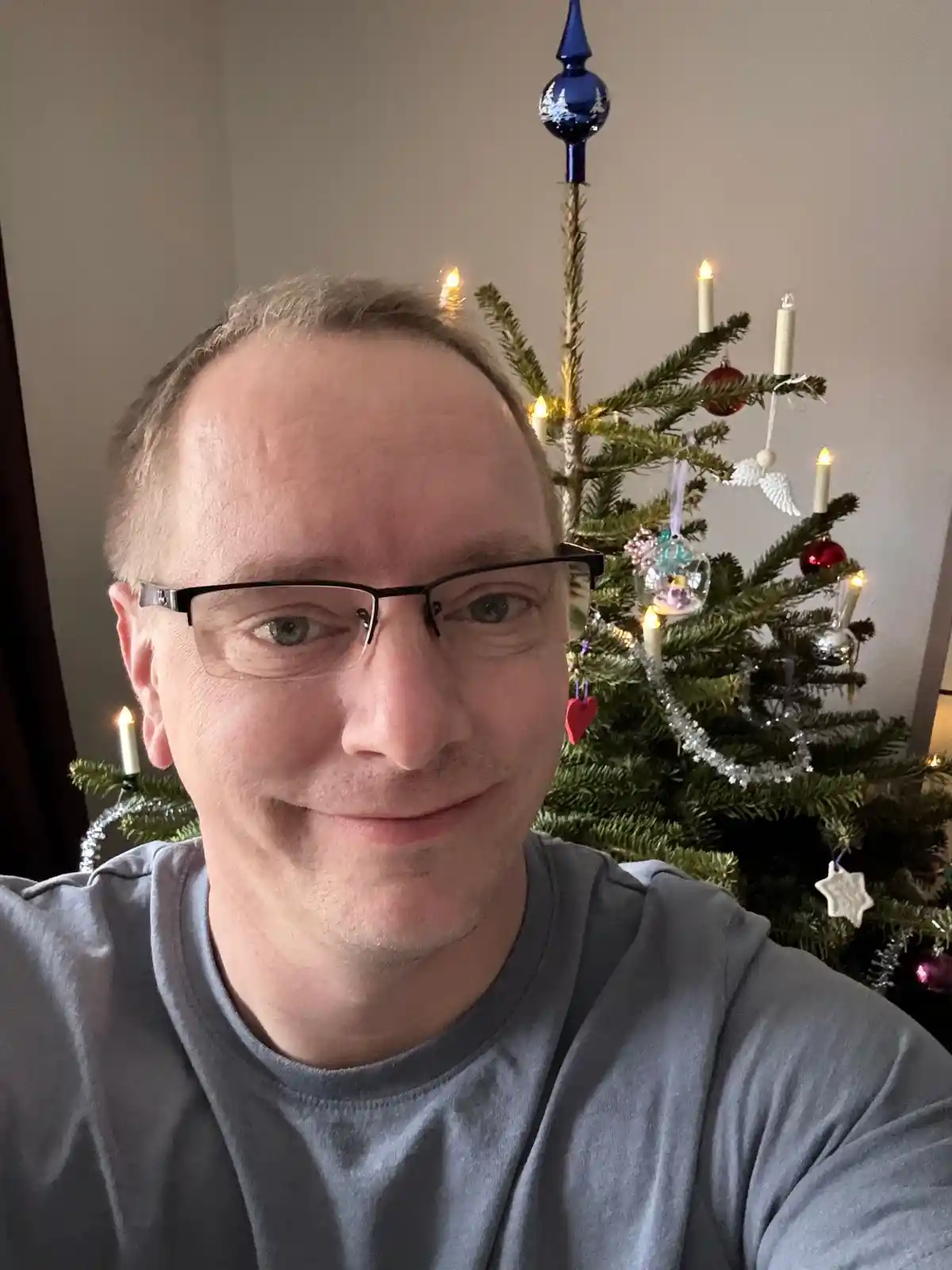 A man with glasses and a slight smirk poses for a selfie in front of a decorated christmas tree with lights, ornaments, and…