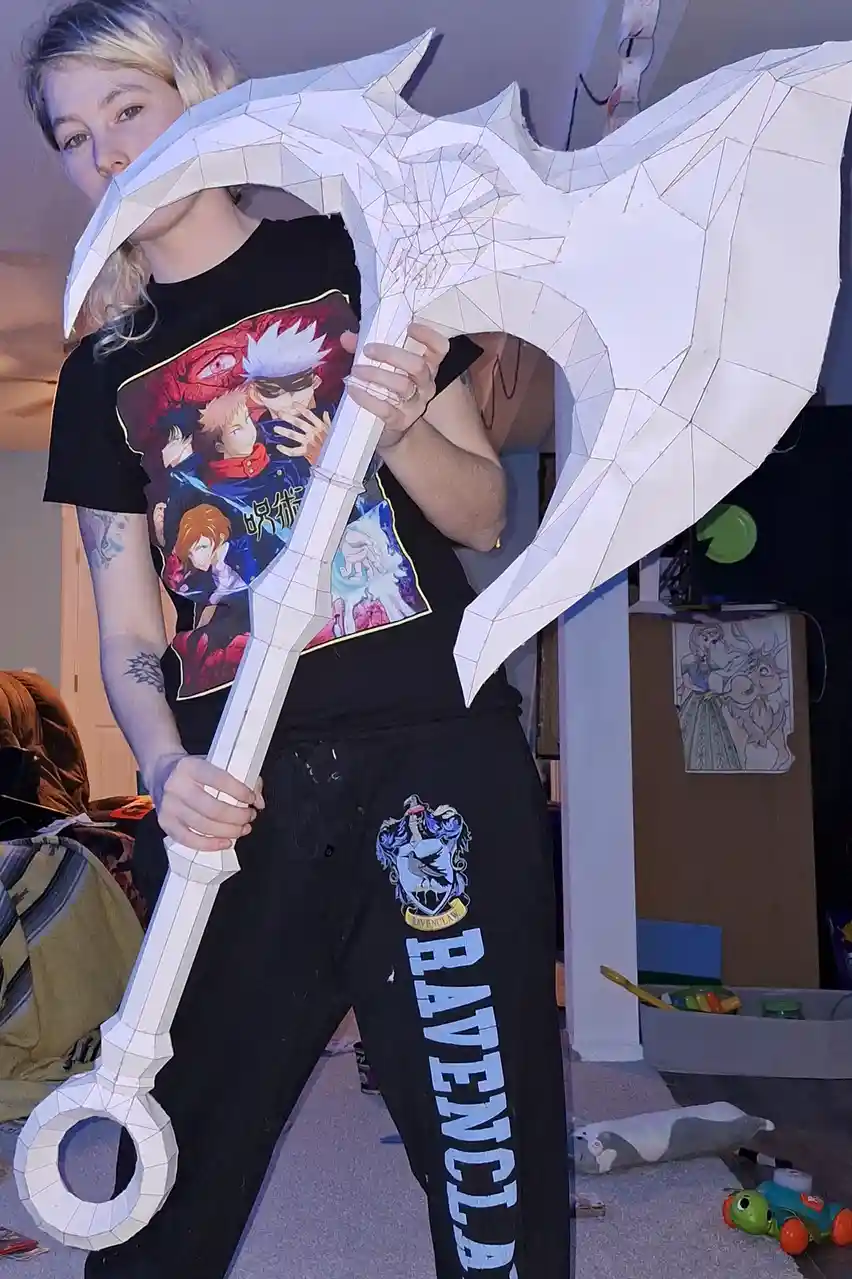A person with blonde hair and tattoos holds a large, white, geometrically faceted paper craft replica of a fantasy-style axe…