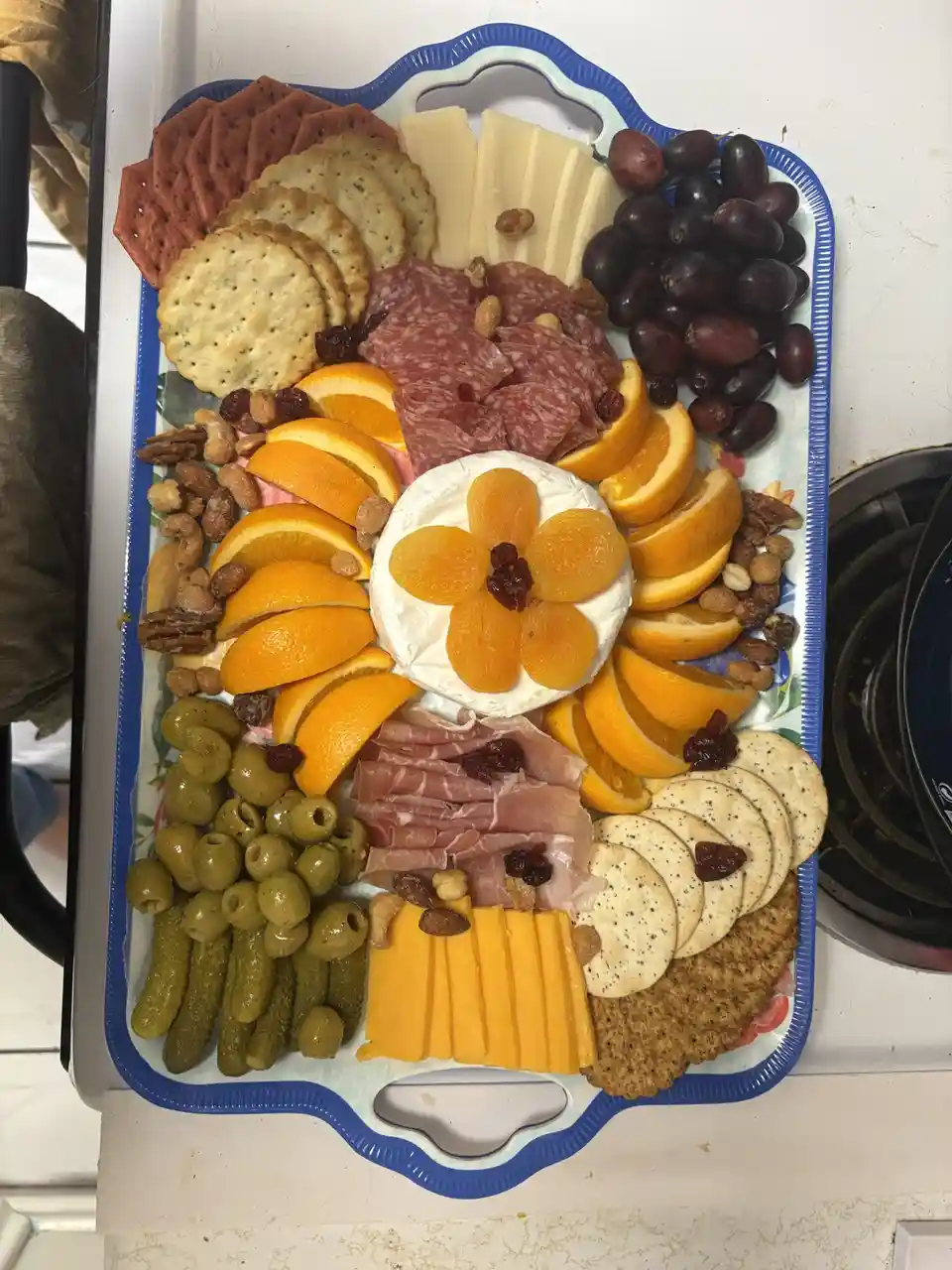 An overhead photo of a colorful, meticulously arranged charcuterie board on a blue-trimmed tray, featuring sliced cheese…