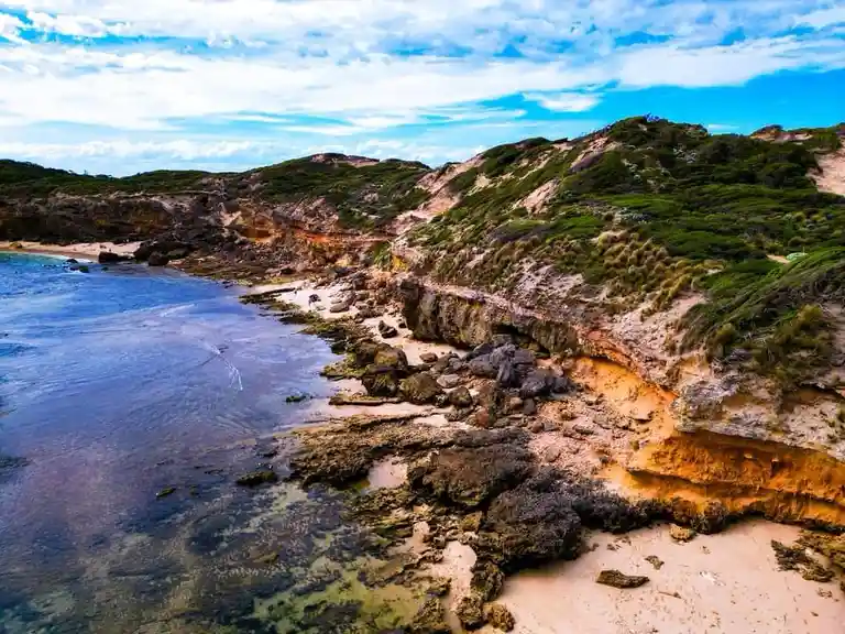 A scenic coastal landscape with sandy beaches, rocky outcrops, and green vegetation under a partly cloudy blue sky. Overlaid…