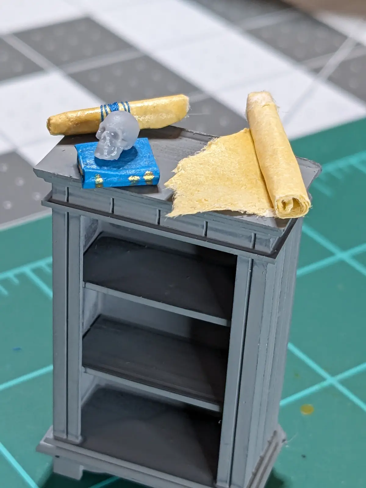A miniature grey bookshelf with an empty interior, topped with a tiny blue book, a translucent skull, a rolled-up scroll…