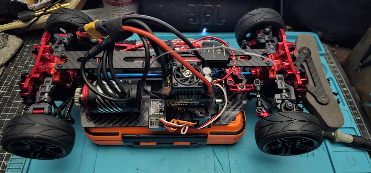 A top-down view of a red and black remote-controlled car chassis with exposed wiring, motor, and electronics, mounted inside…