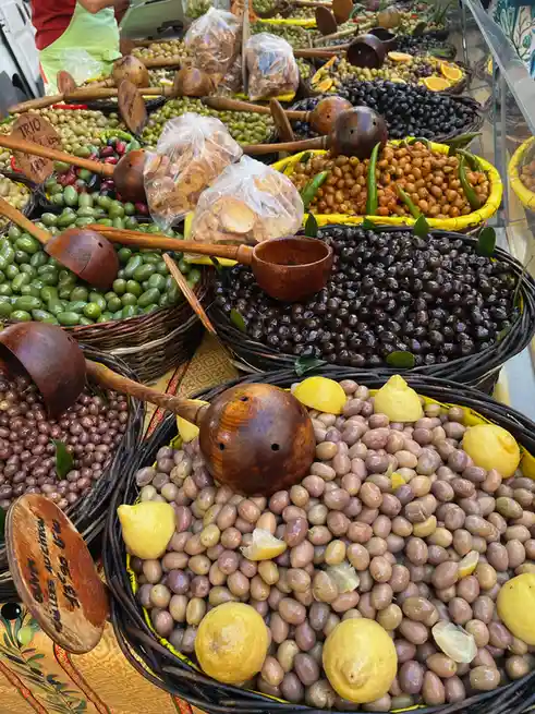 A vibrant market stall displays numerous woven baskets filled with various types of olives—green, black, and purple—some…