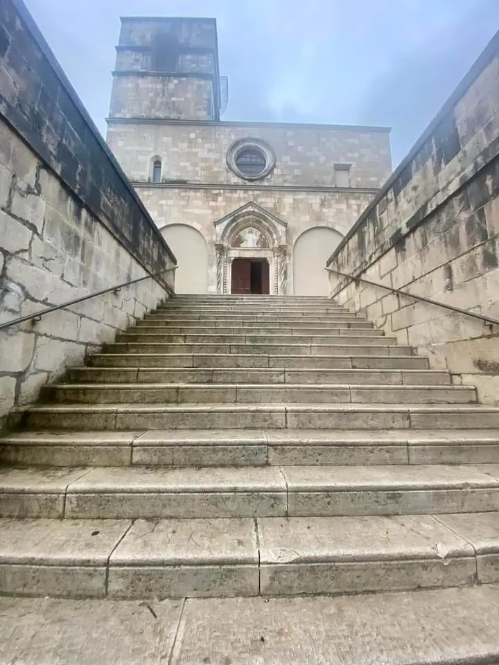 A low-angle photo shows a long, wide stone staircase leading up to a large, historic church building with a bell tower and…