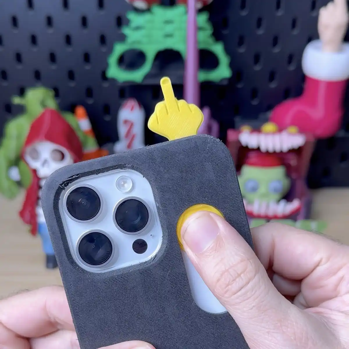 A hand holds a smartphone with a black case, showing a yellow 3d-printed finger pointing up from the top of the case. The…