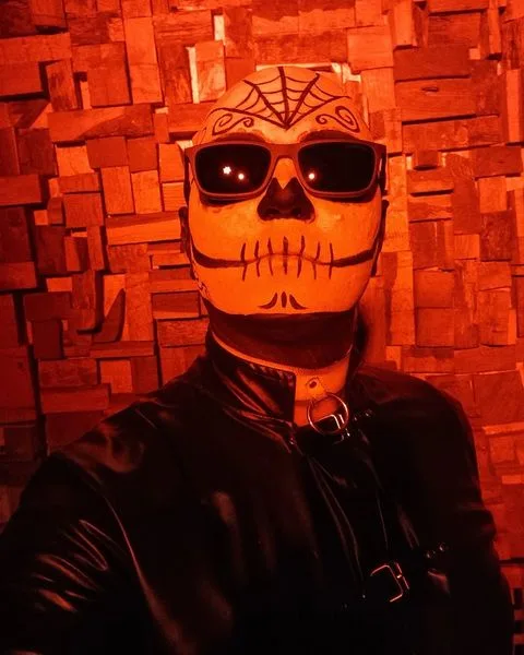 A person wearing a skull mask with decorative swirls and a spiderweb pattern, dark sunglasses, and a black leather jacket…