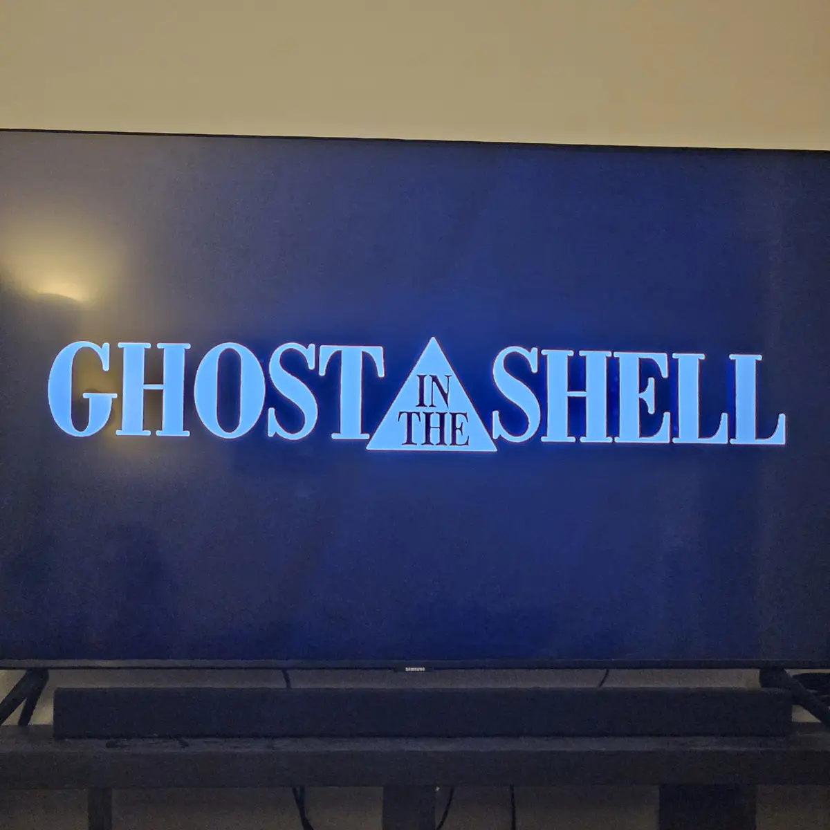 A television screen displays the title 'GHOST IN THE SHELL' in glowing blue letters, with the word 'IN' replaced by a…
