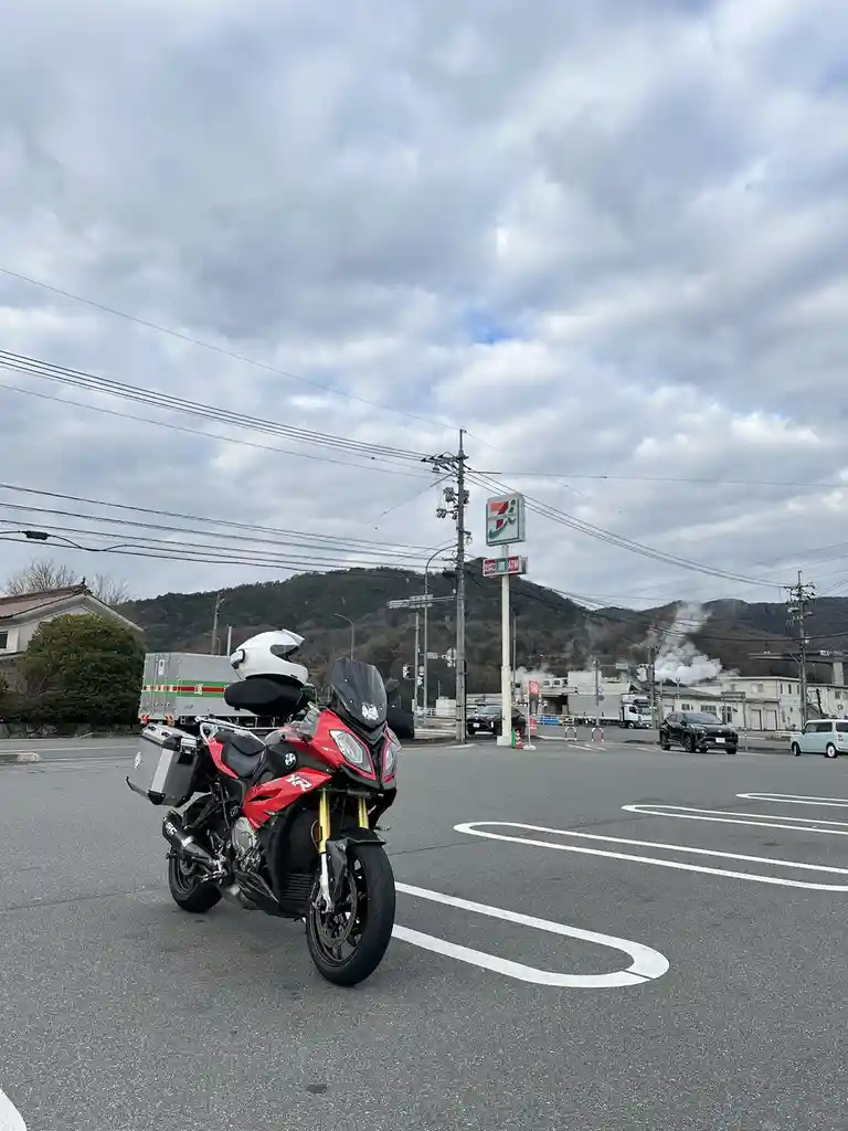 A red and black BMW motorcycle with a white helmet on its top case is parked in a paved lot under a cloudy sky. In the…