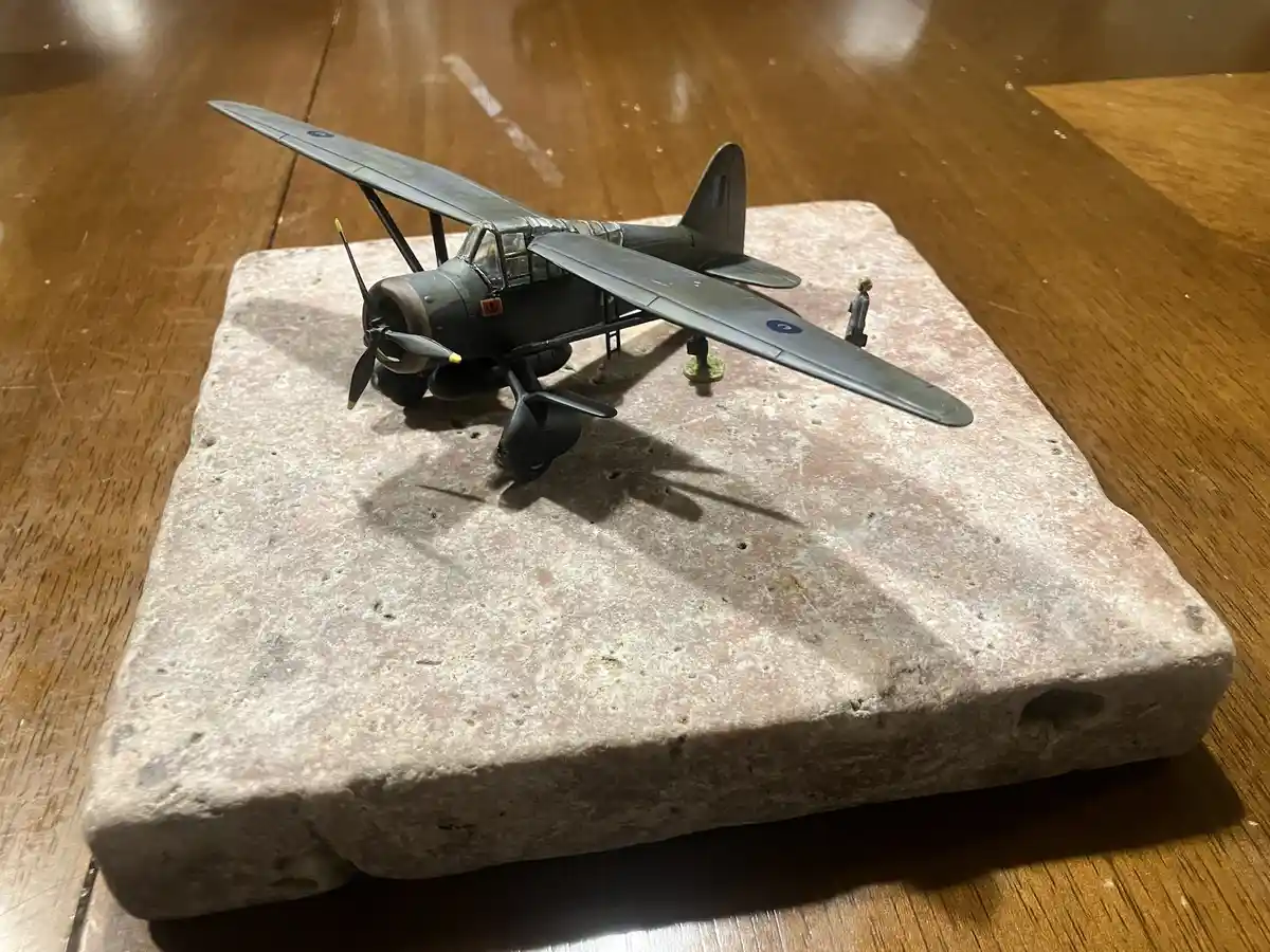 A small, detailed model of a grey biplane with blue roundels sits atop a large, rough-hewn rectangular stone block, which…