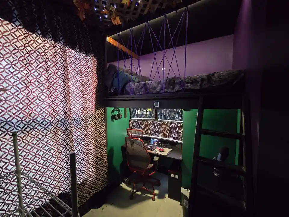 A dimly lit, eclectic room features a loft bed with purple ropes and blankets, above a green-walled desk with a red gaming…