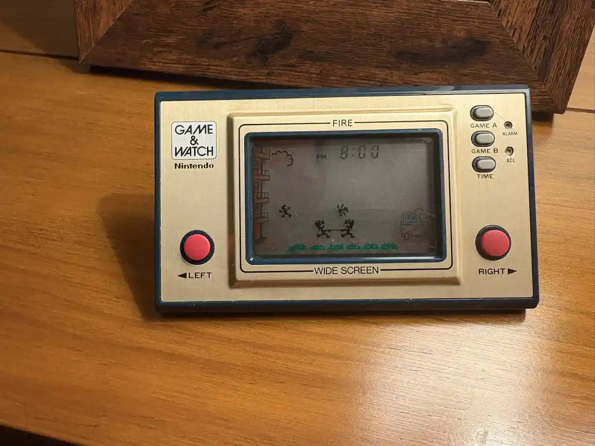 A vintage Nintendo Game & Watch device, specifically the 'Fire' model, rests on a wooden surface. Its screen displays a…