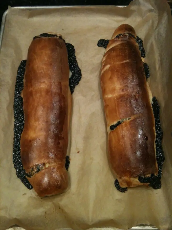 Two golden-brown baked rolls sit on parchment paper on a baking tray, each with a dark, black, licorice-like substance…