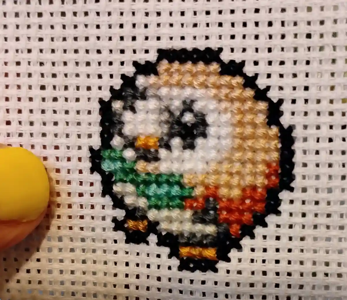 A close-up of a cross-stitch pattern depicting a pixelated character with a black outline, white face, brown hair, green…