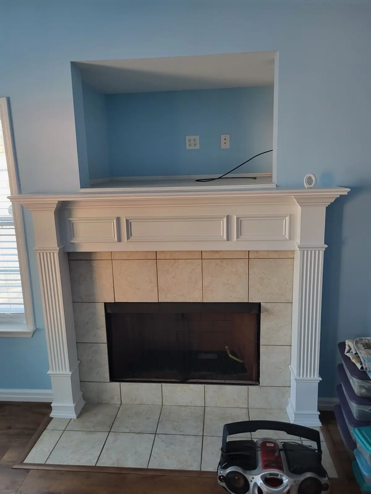 A white fireplace with beige tiled surround and mantel sits in a blue-walled room; a boombox rests on the tiled floor in…