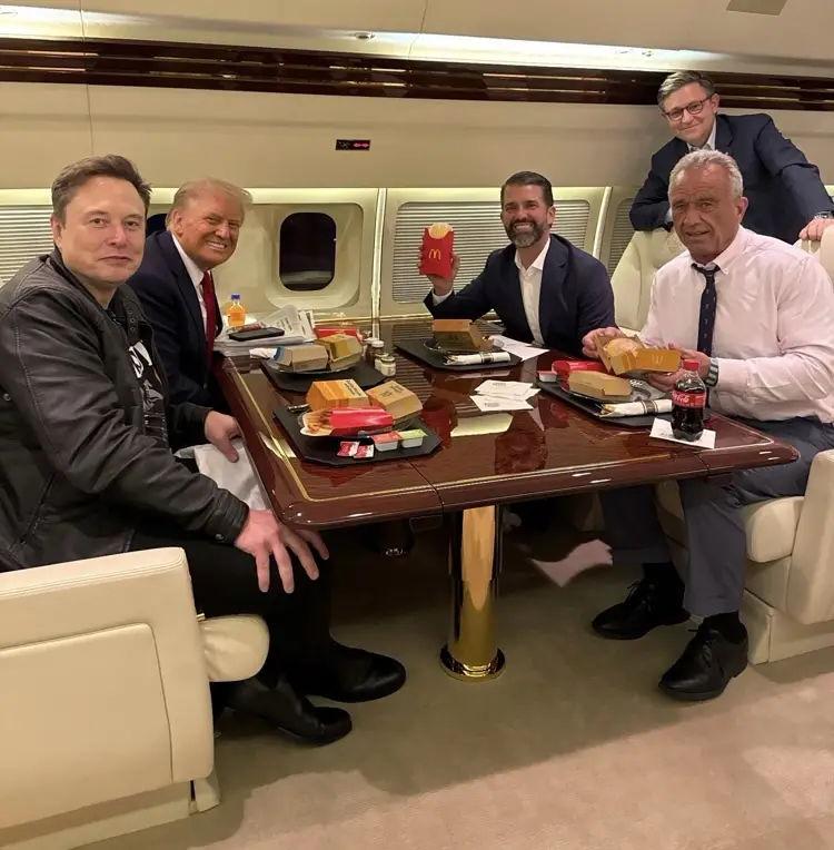 Five men are seated around a polished wooden table inside a private jet cabin, eating McDonald's meals. Elon Musk, Donald…