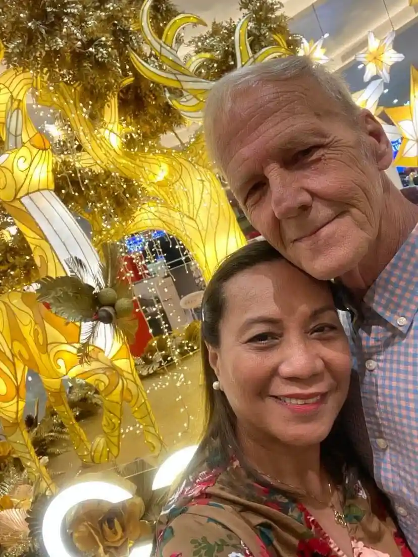 An older man and woman pose for a selfie in front of a large, ornate golden reindeer decoration with glowing lights. The man…