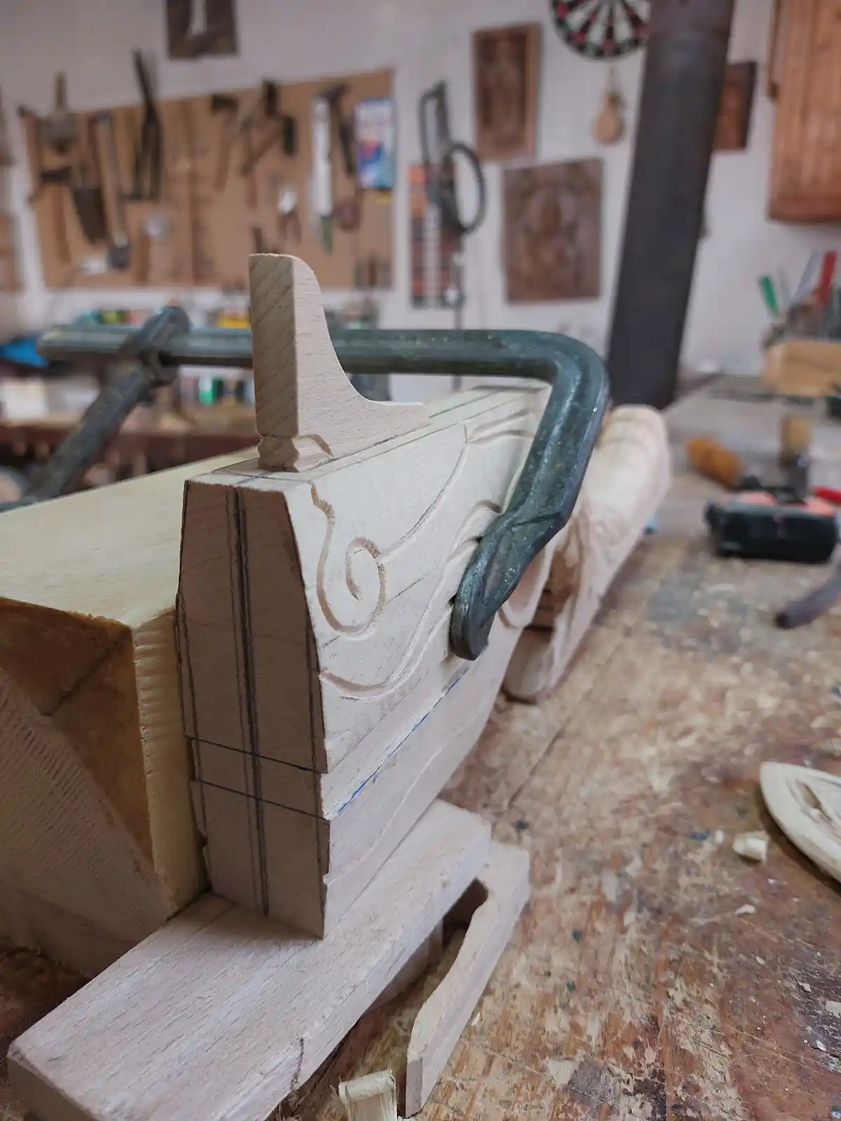 A close-up photo of a wooden carving in progress, held in a metal clamp on a workbench in a cluttered woodshop. The carving…