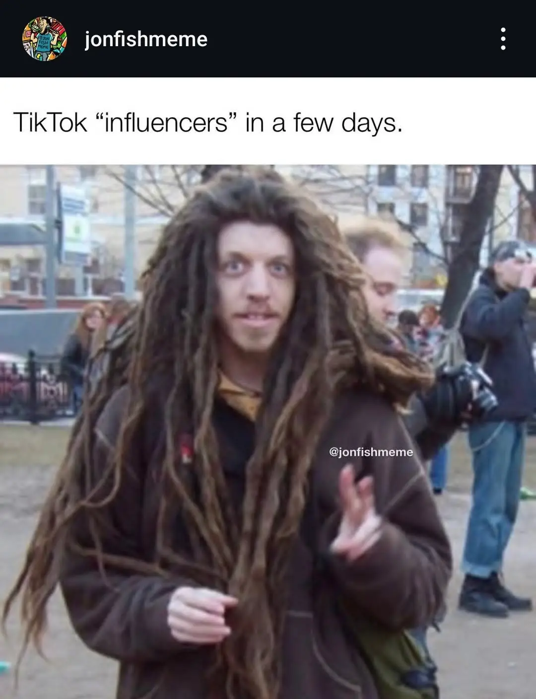 A man with long, messy dreadlocks and a brown jacket stands outdoors, looking directly at the camera with a slightly…