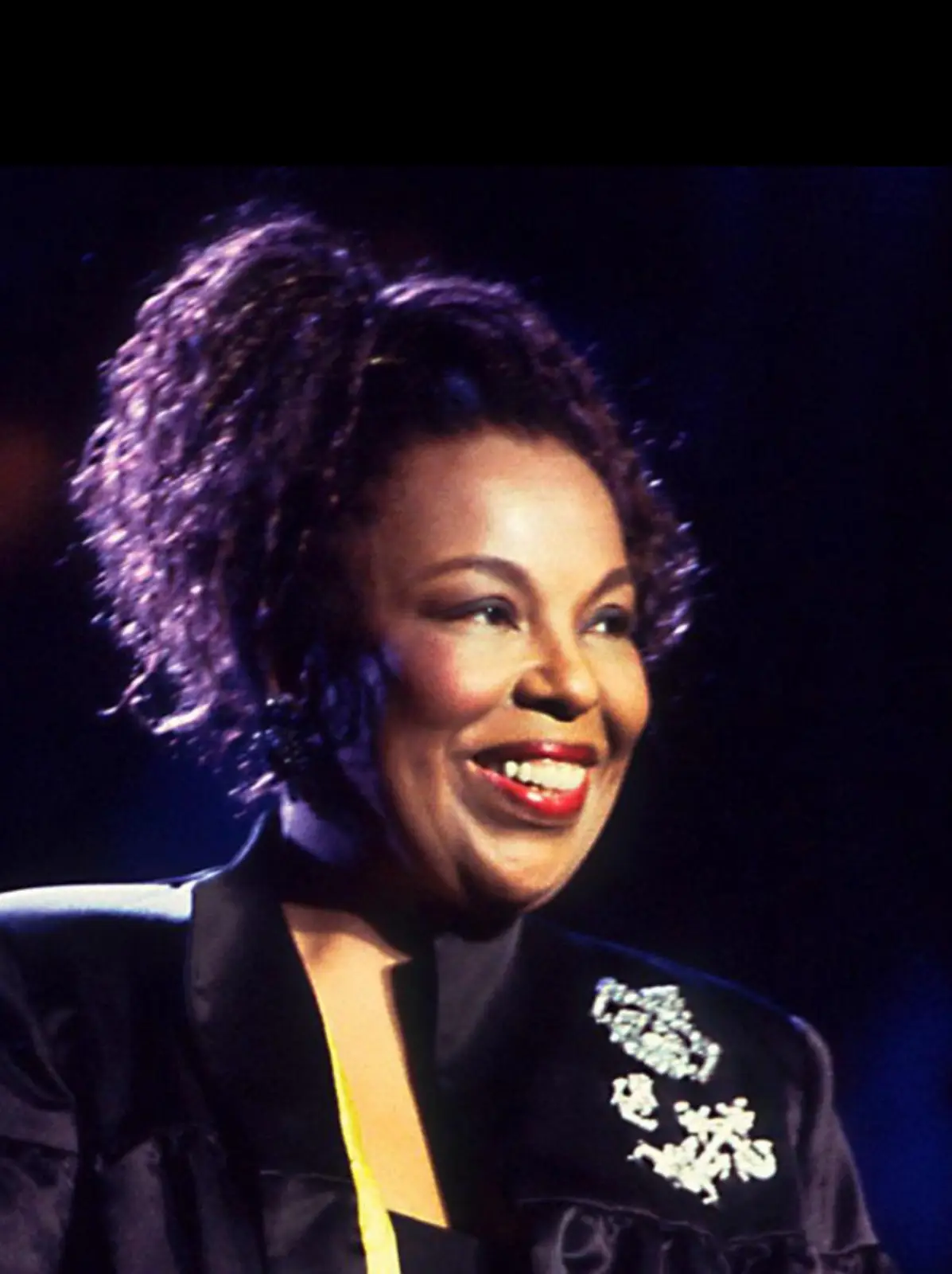 a close-up photo of a smiling woman with curly hair, wearing a black jacket with white embroidery, against a dark…