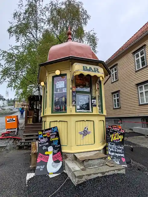 A small, yellow, dome-topped bar kiosk with a sign reading 'BAR' and 'OPEN ALL DAYS 12-19'. Two chalkboards flank the…