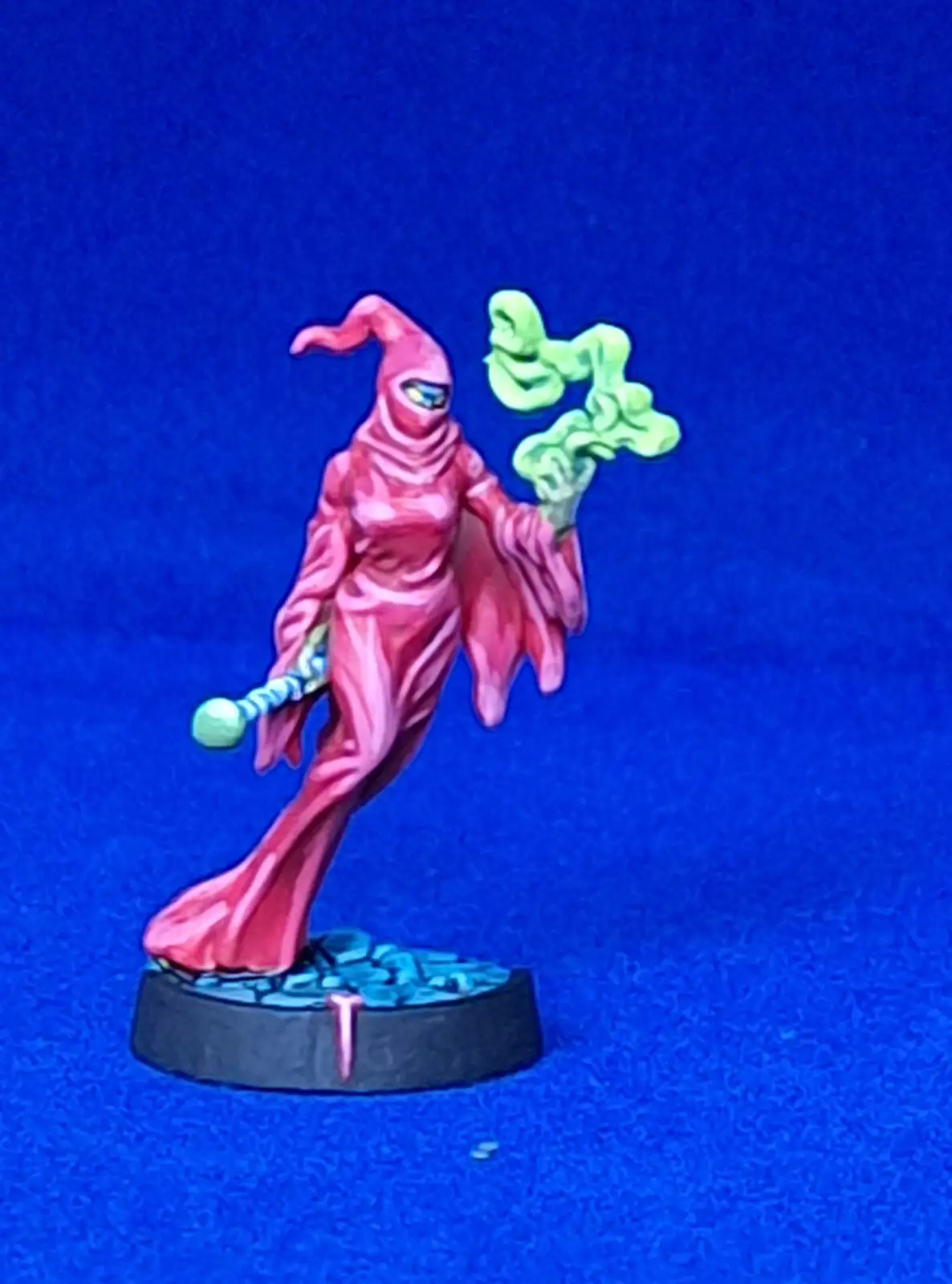 A painted miniature of a pink-robed, hooded figure resembling a witch or wizard stands on a circular base against a solid…