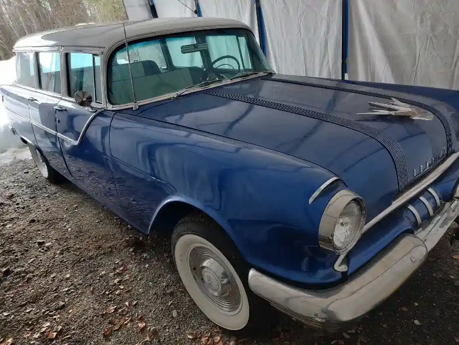 A blue 1950s Chevrolet station wagon with chrome trim and whitewall tires is parked on gravel near a white tarp. The car has…