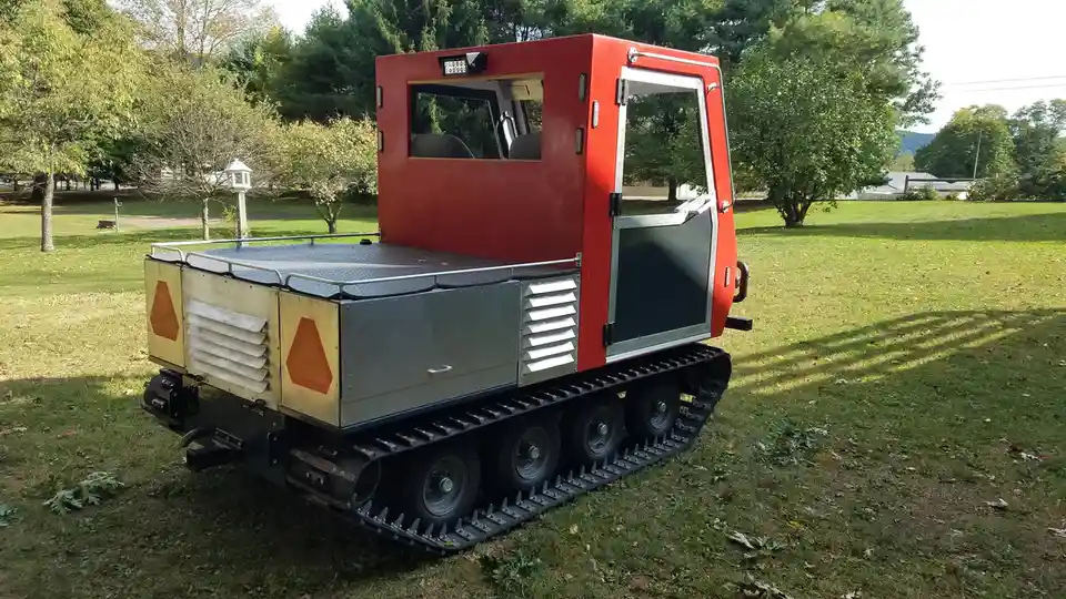 A red and gray tracked vehicle with a boxy cab and orange hazard markings sits on grass, resembling a tank but with a…
