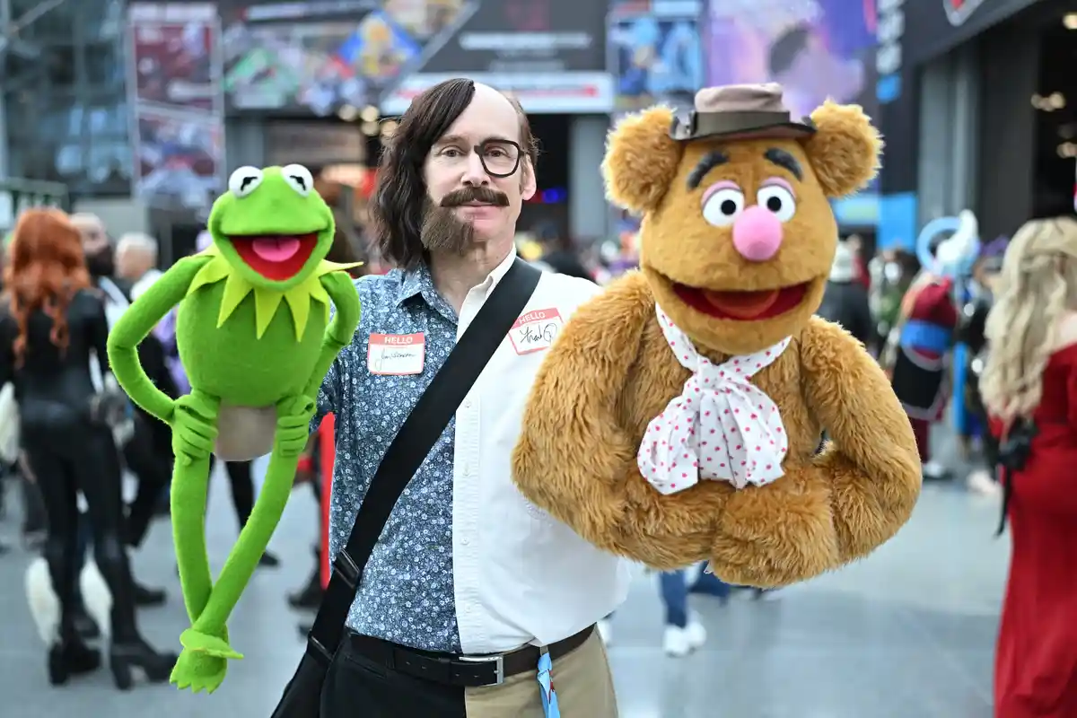 A man with a mustache and glasses, wearing a name tag that reads 'hello my name is kermit the frog', stands between two…