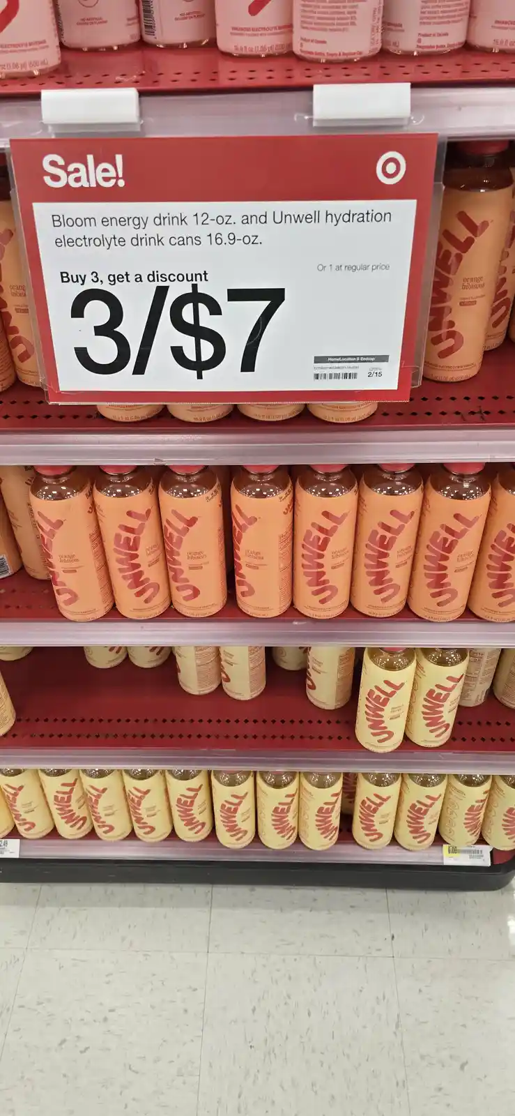 A Target store shelf displays rows of Unwell electrolyte drink cans in orange and yellow packaging. A red sale sign above…