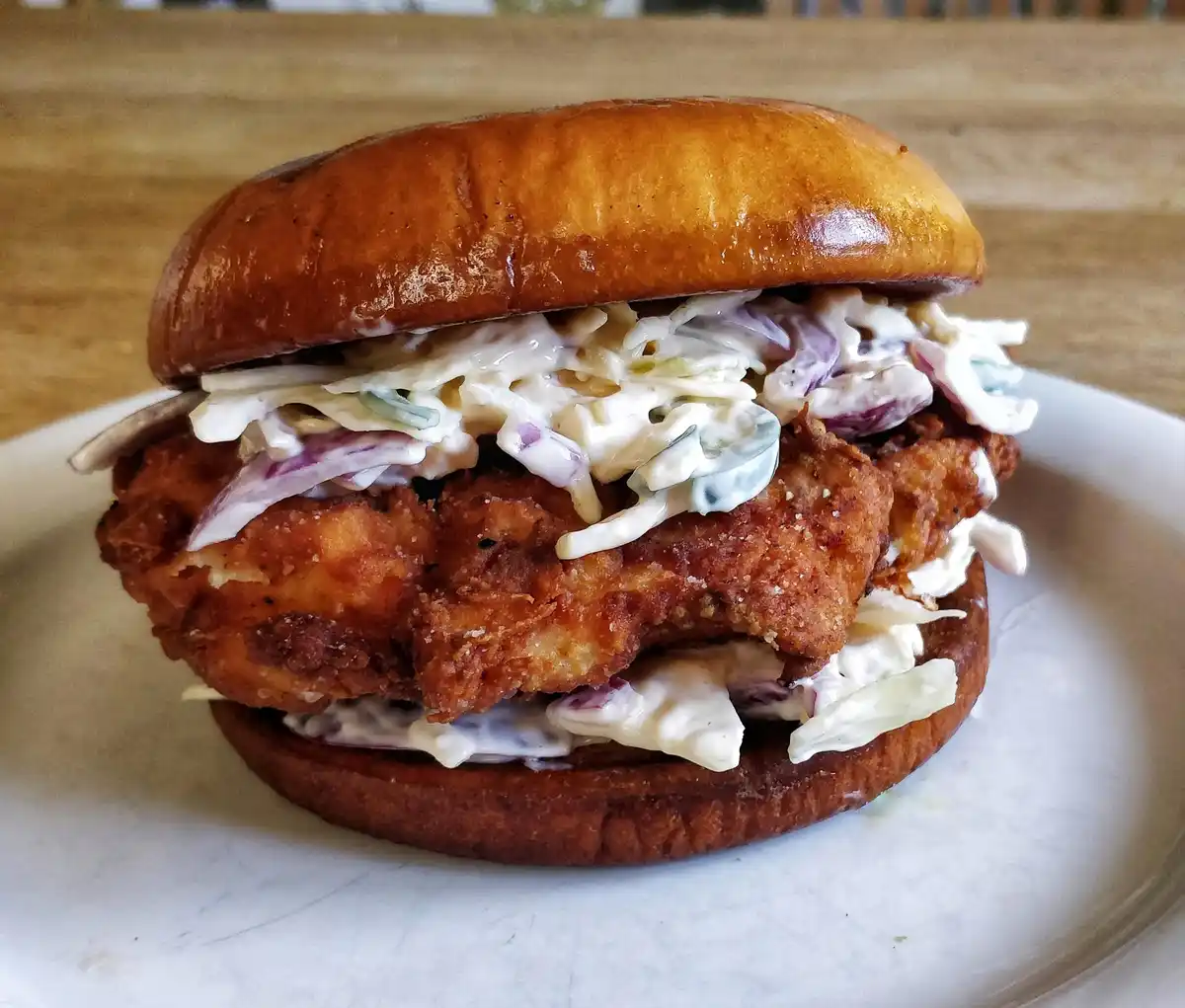 A close-up photo shows a fried chicken sandwich on a pretzel-style bun, generously topped with creamy coleslaw containing…
