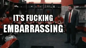 A man in a suit and red tie stands in a locker room with players in orange uniforms seated behind him. He initially stands…